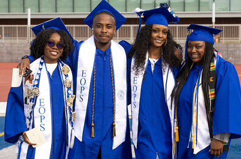 Four graduates in blue caps and gowns stand together on an outdoor track with arms around each other. Each graduate wears a white EOPS stole, with additional cords and decorations visible on some stoles.