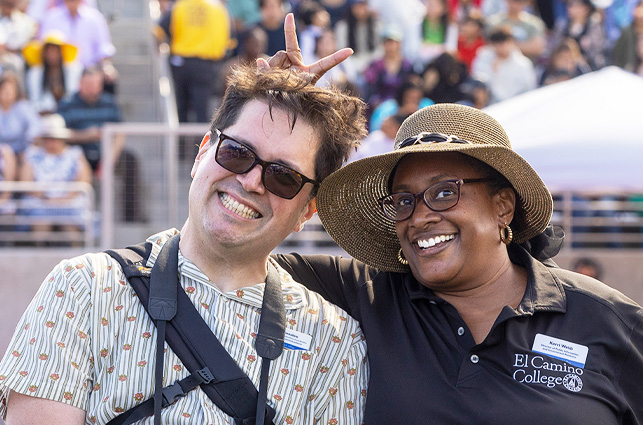Two individuals standing close together at an outdoor event. One person is making a playful hand gesture above the other person’s head. The individual on the right is wearing a wide-brimmed hat and a dark El Camino College polo shirt with a visible name badge. The background shows a crowd of people seated in bleachers and event tents.
