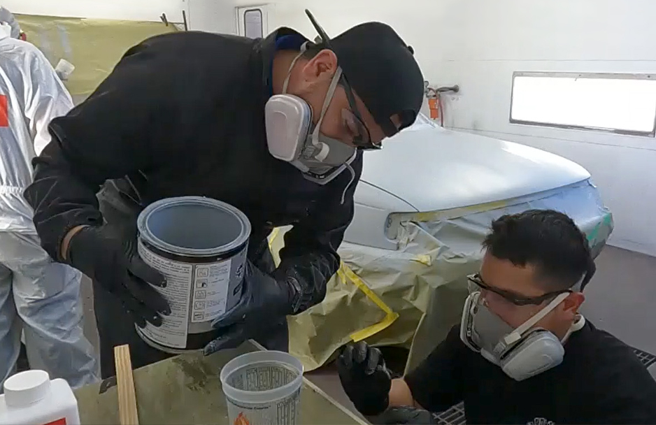 Two people in respirators mix paint in an auto body shop beside a masked car.