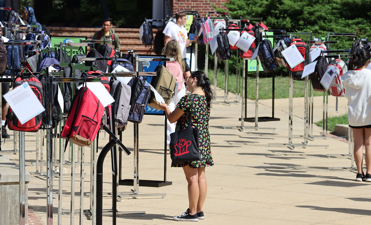 Rows of backpacks hang on metal stands outdoors, each with a paper attached, at a Send Silence Packing exhibit. People walk among them reading the papers, with trees and a brick building in the background.